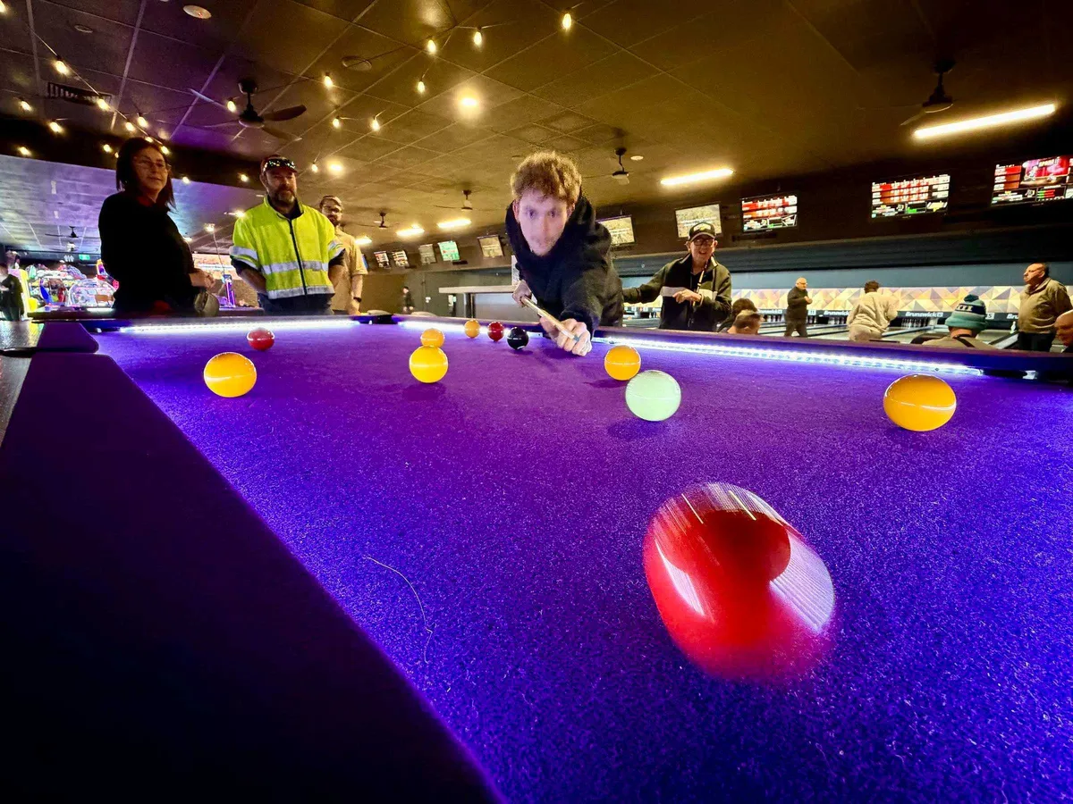 Friends watching a shot on the purple-felt pool table, with the bowling lanes behind, at Bendigo Bowl